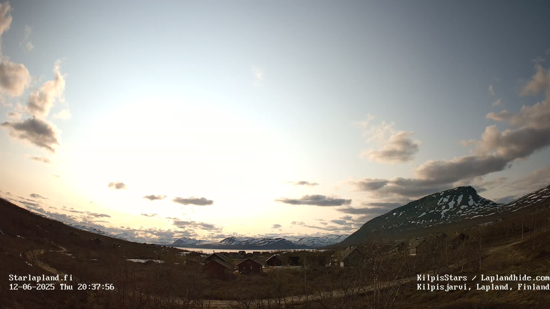 A partly cloudy sunset over a mountainous landscape with a small village nestled among sparse trees.