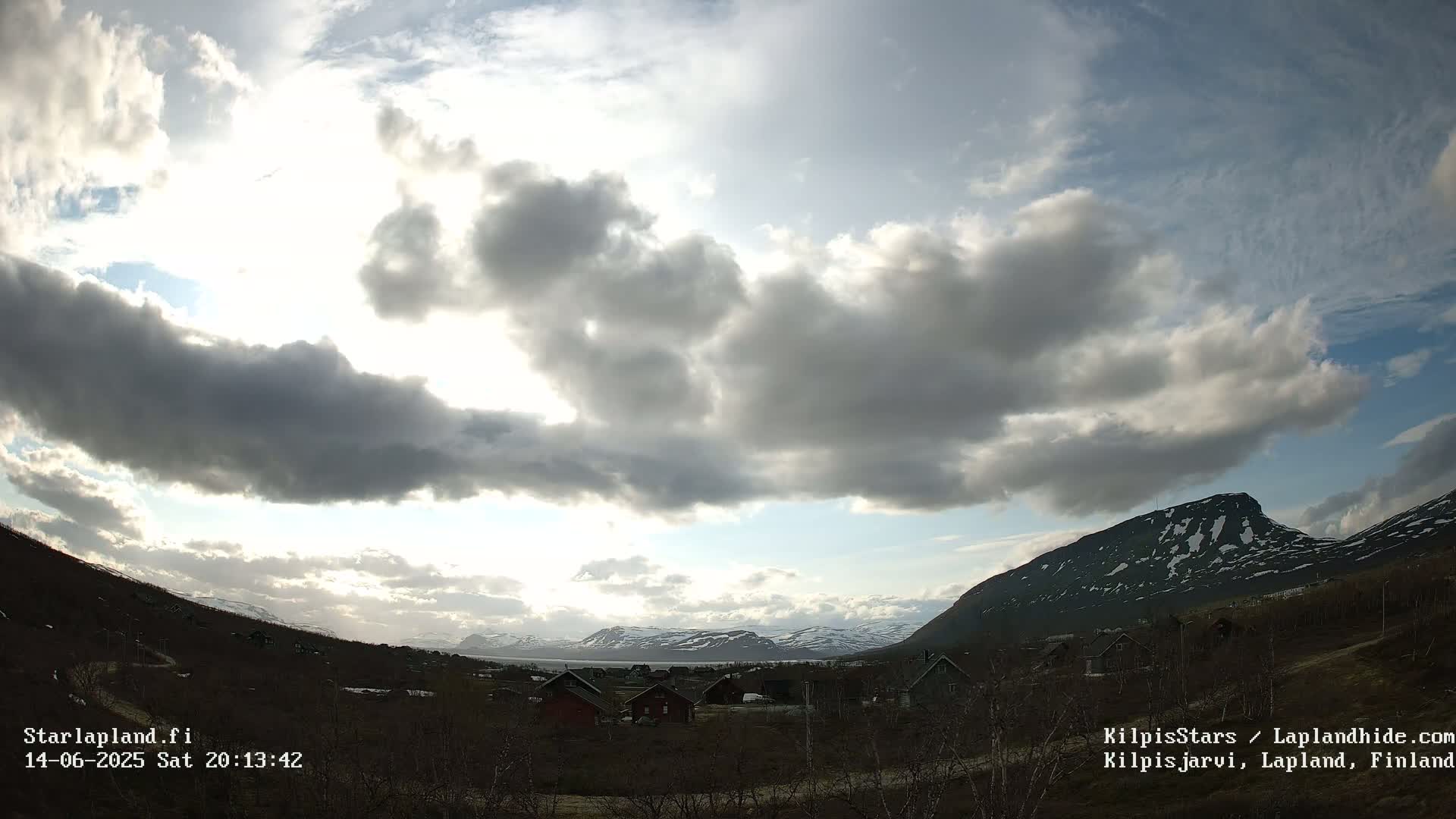 A partly cloudy sky brightens over a valley of small houses nestled between low, snow-capped mountains.