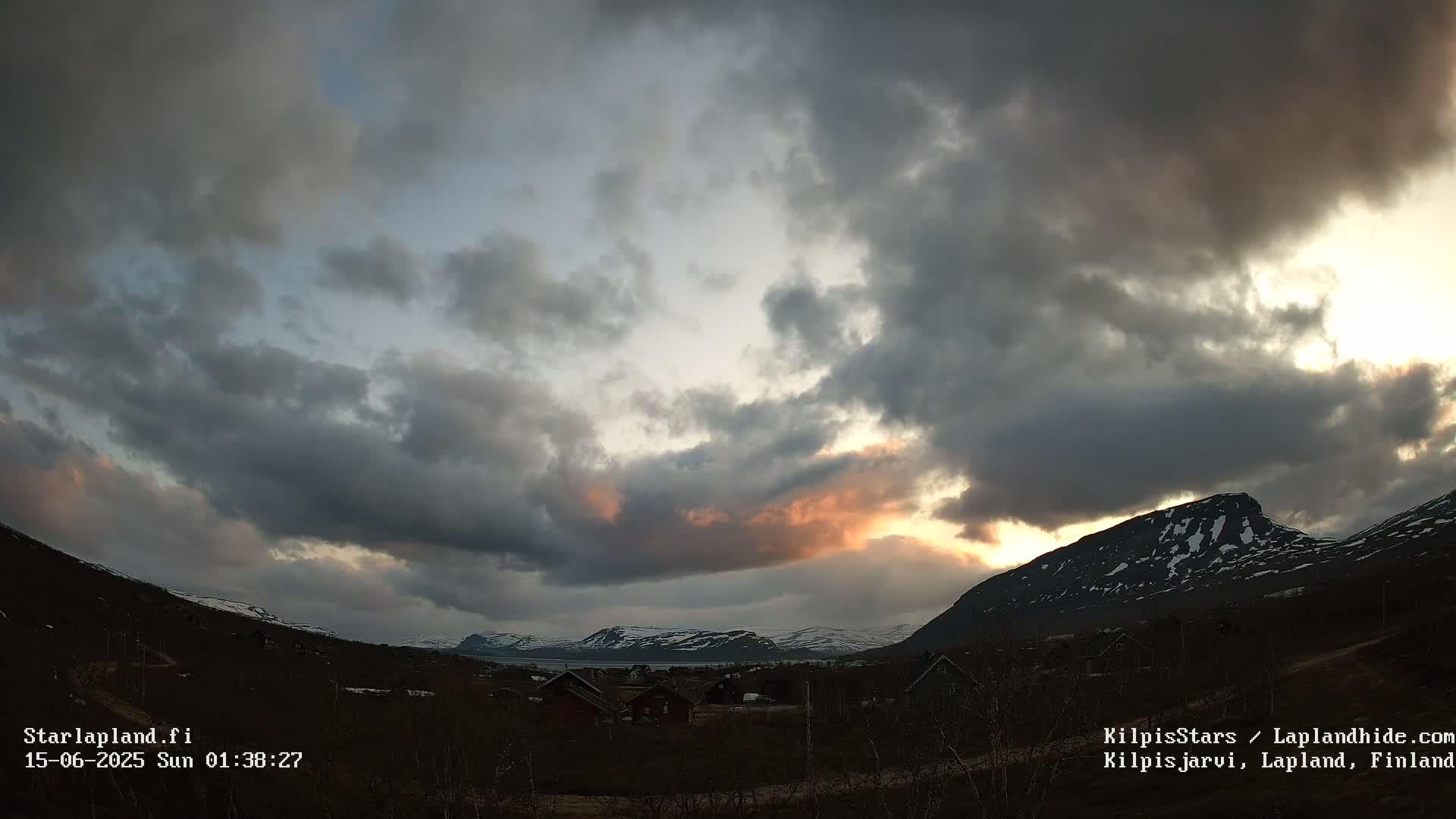 A partly cloudy sky brightens over a valley of small houses nestled between low, snow-capped mountains.