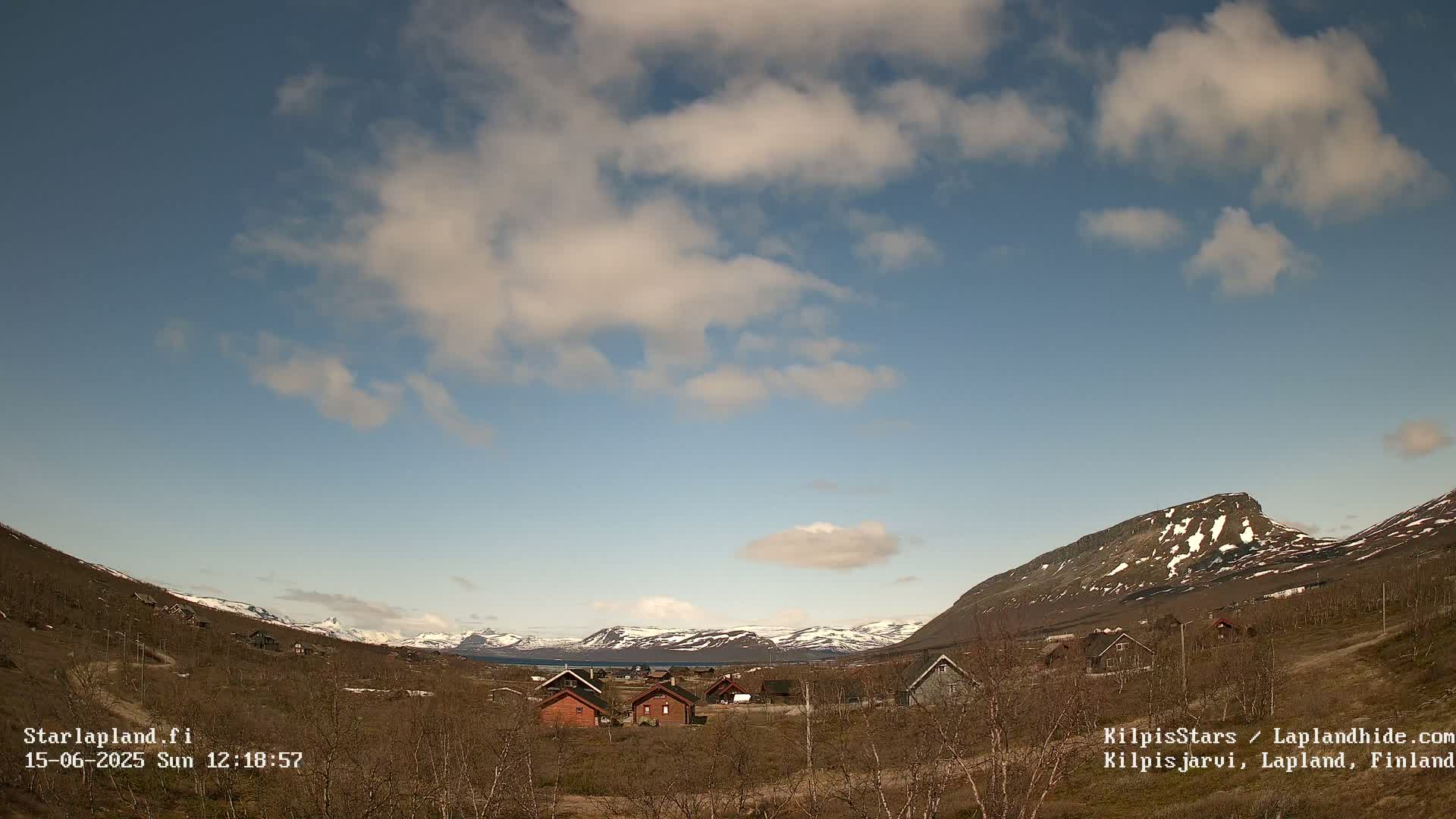 A partly cloudy sky overlooks a small cluster of houses nestled in a valley between snow-capped mountains.