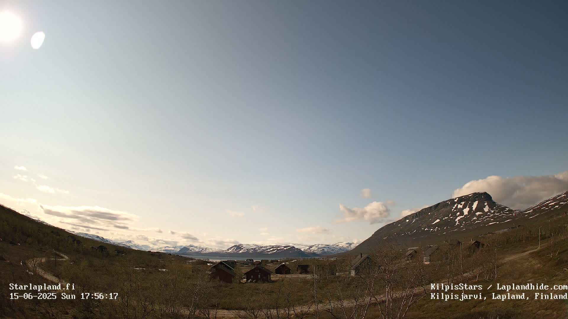 A sunny day with partly cloudy skies shows a small village nestled in a valley between snow-capped mountains.