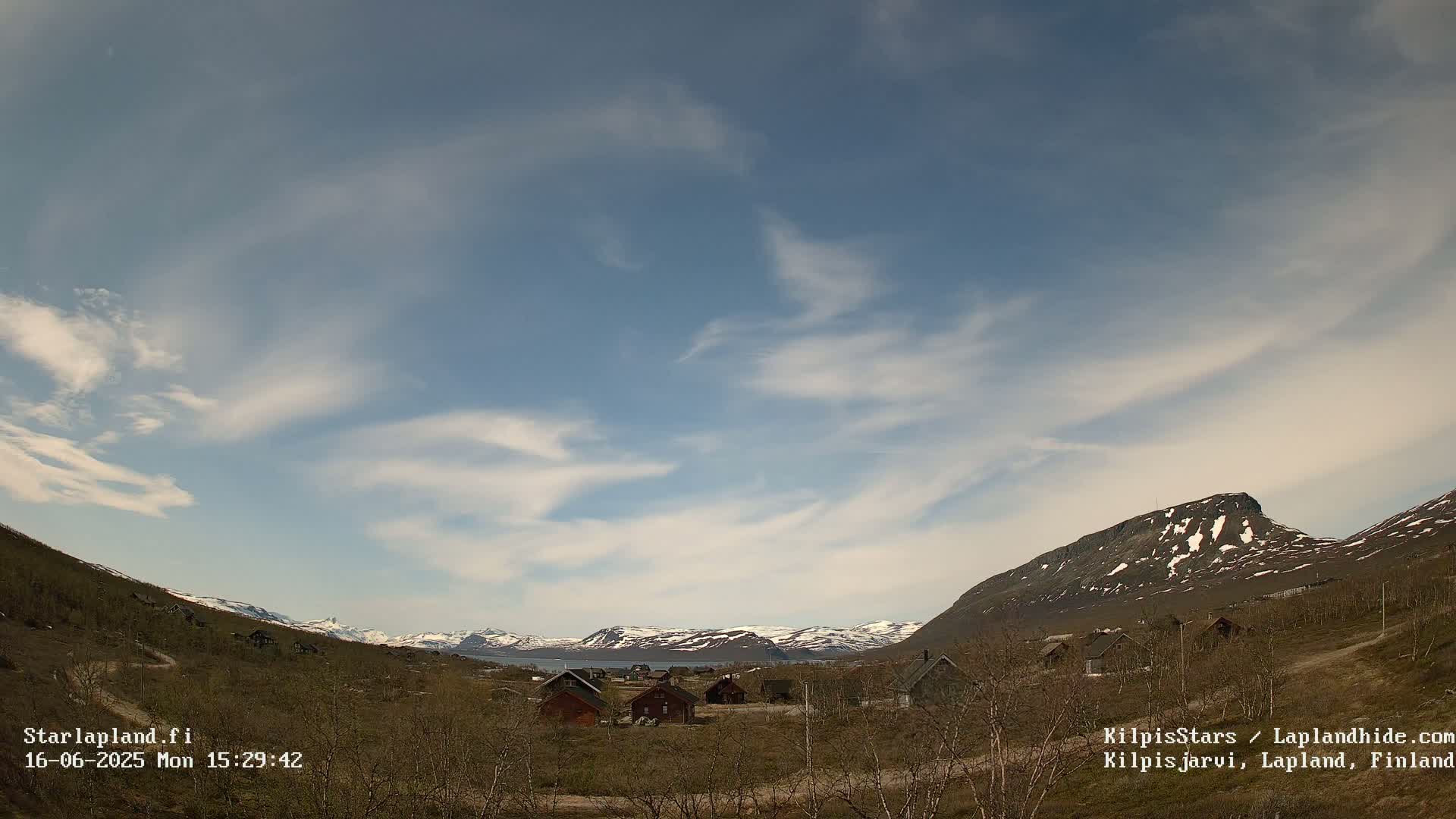 A cluster of small buildings sits in a valley between snow-capped mountains under a mostly clear sky with wispy clouds.