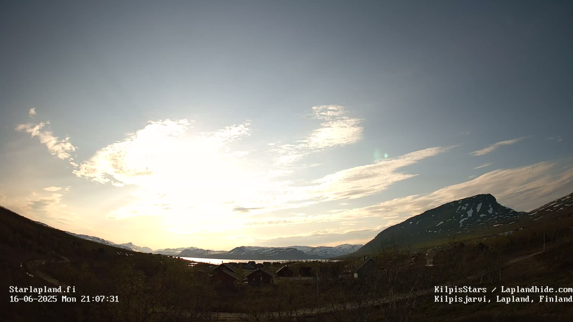A partly cloudy sunset illuminates a village nestled between a lake and snow-capped mountains.