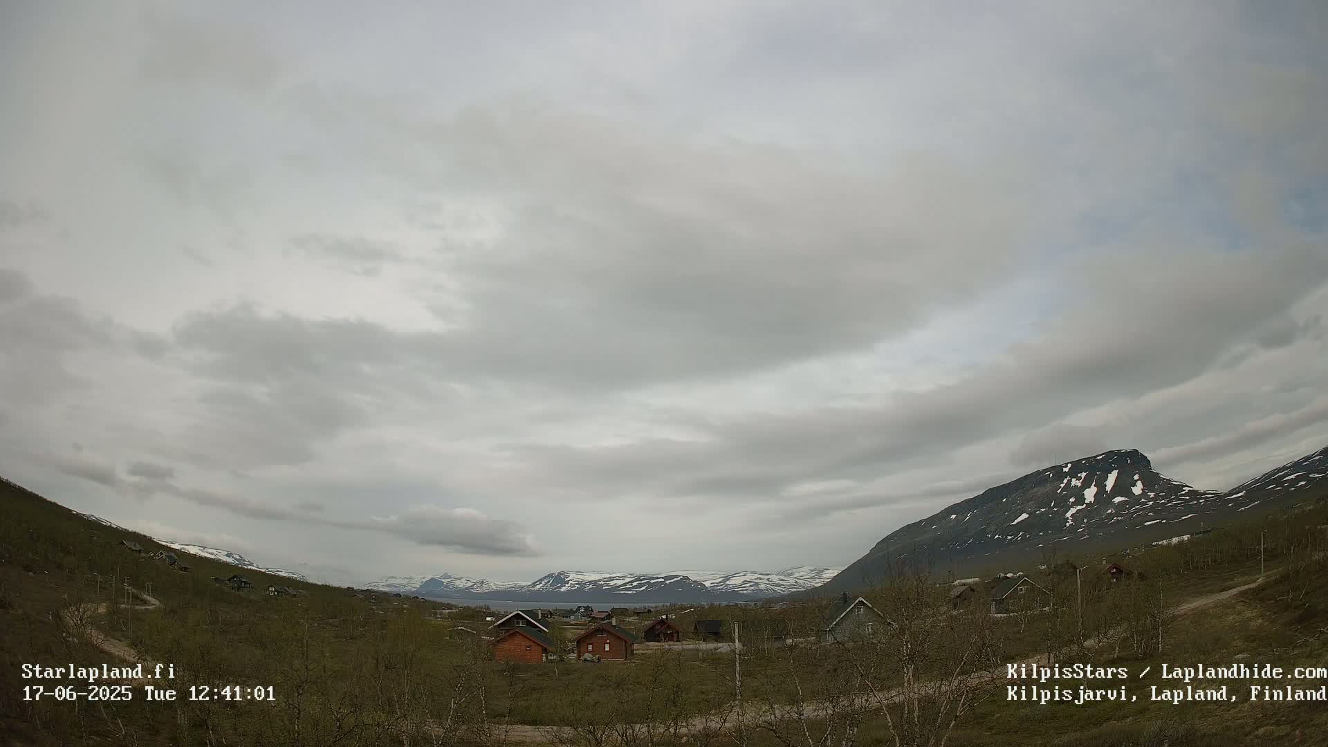 A small cluster of cabins sits in a valley between snow-capped mountains under an overcast sky.