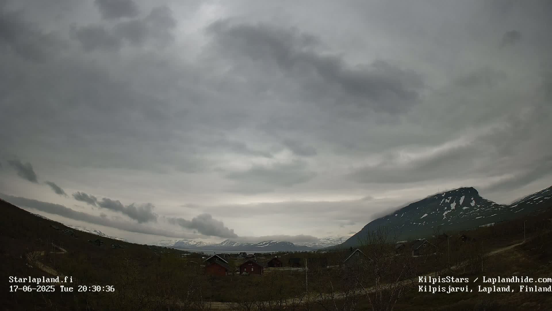 A cloudy, overcast sky hangs over a small village nestled in a valley between snow-dusted mountains.