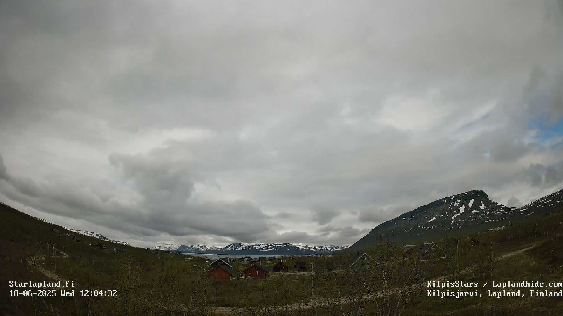 A mostly cloudy sky over a valley of small houses, sparse vegetation, and snow-capped mountains in the distance.