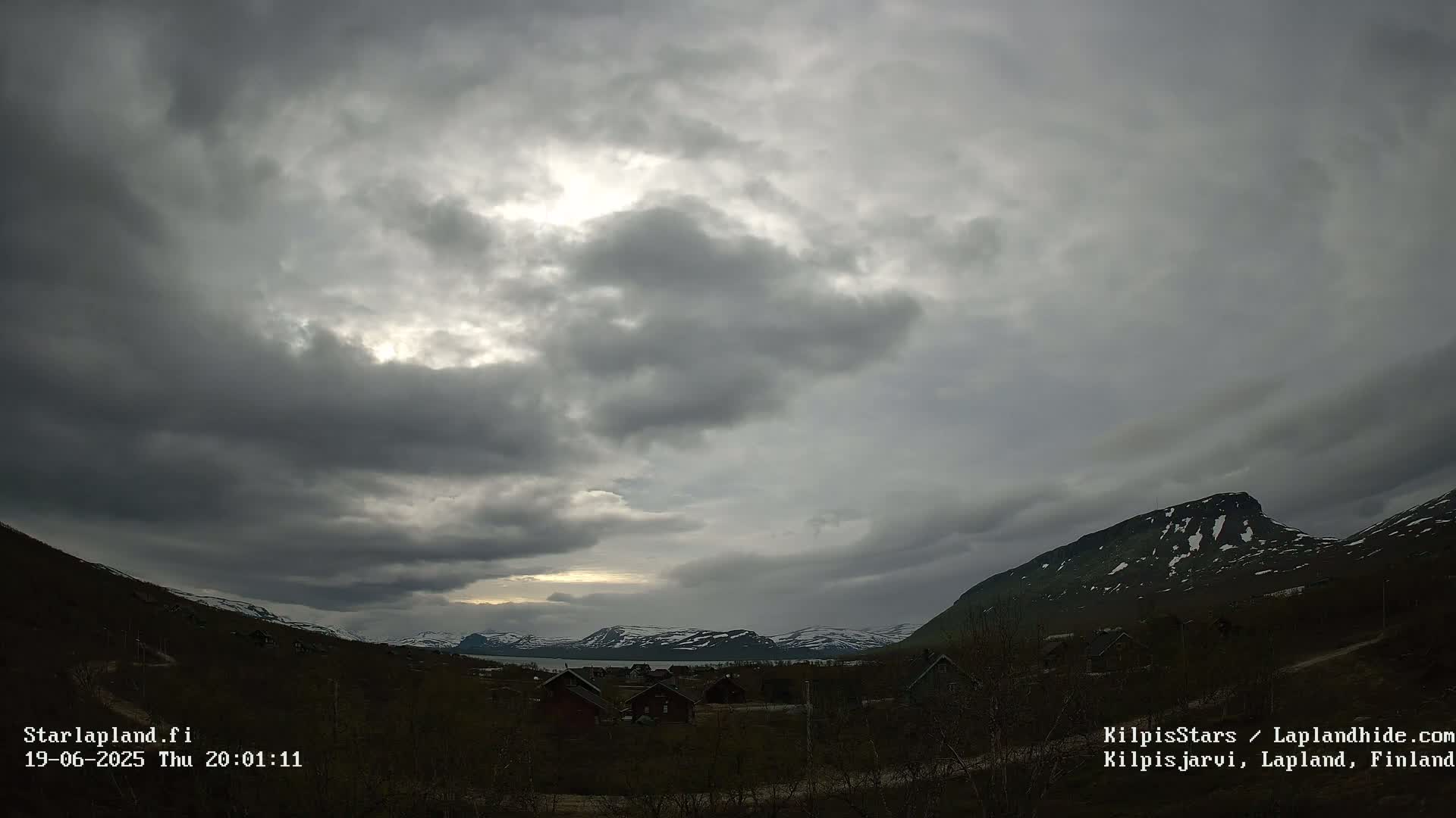 A mostly cloudy sky over a valley with small houses and snow-capped mountains in the distance.