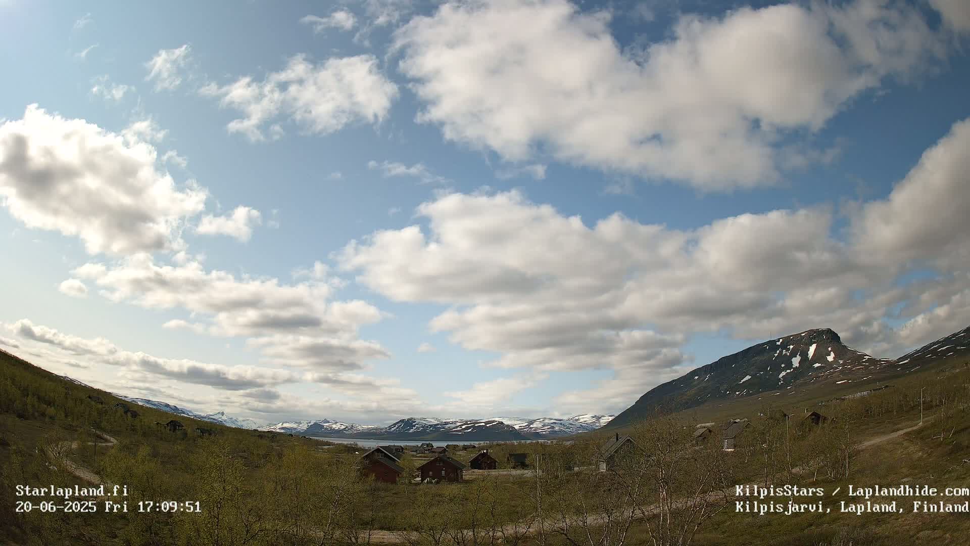 A partly cloudy sky overlooks a valley of sparse, low-lying vegetation with small houses nestled amongst it, backed by distant mountains with some snow patches.
