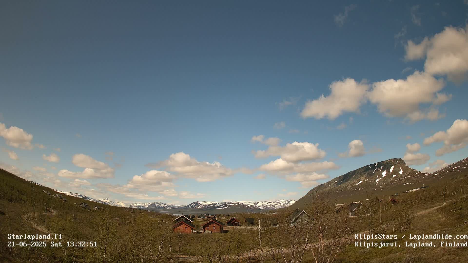 A small cluster of cabins sits in a valley between snow-capped mountains under a partly cloudy sky.