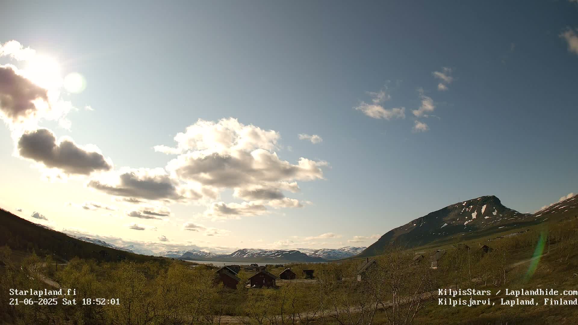 A partly sunny sky overlooks a valley of small cabins nestled amongst low, green vegetation, with snow-capped mountains in the background.