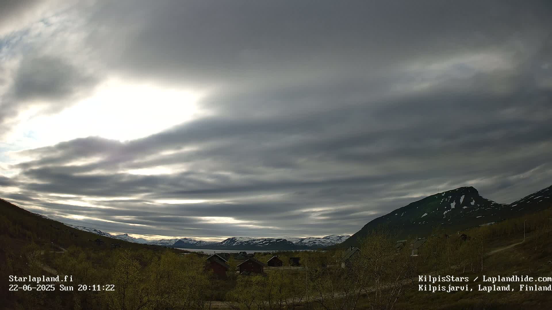 A partly cloudy sky over a mountainous landscape with a few small buildings and sparse, budding trees in the foreground.