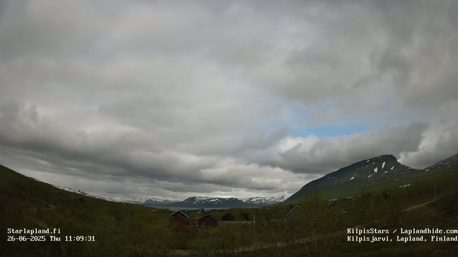 A mostly cloudy sky hangs over a valley of low-lying vegetation and small cabins, with snow-capped mountains in the distance.