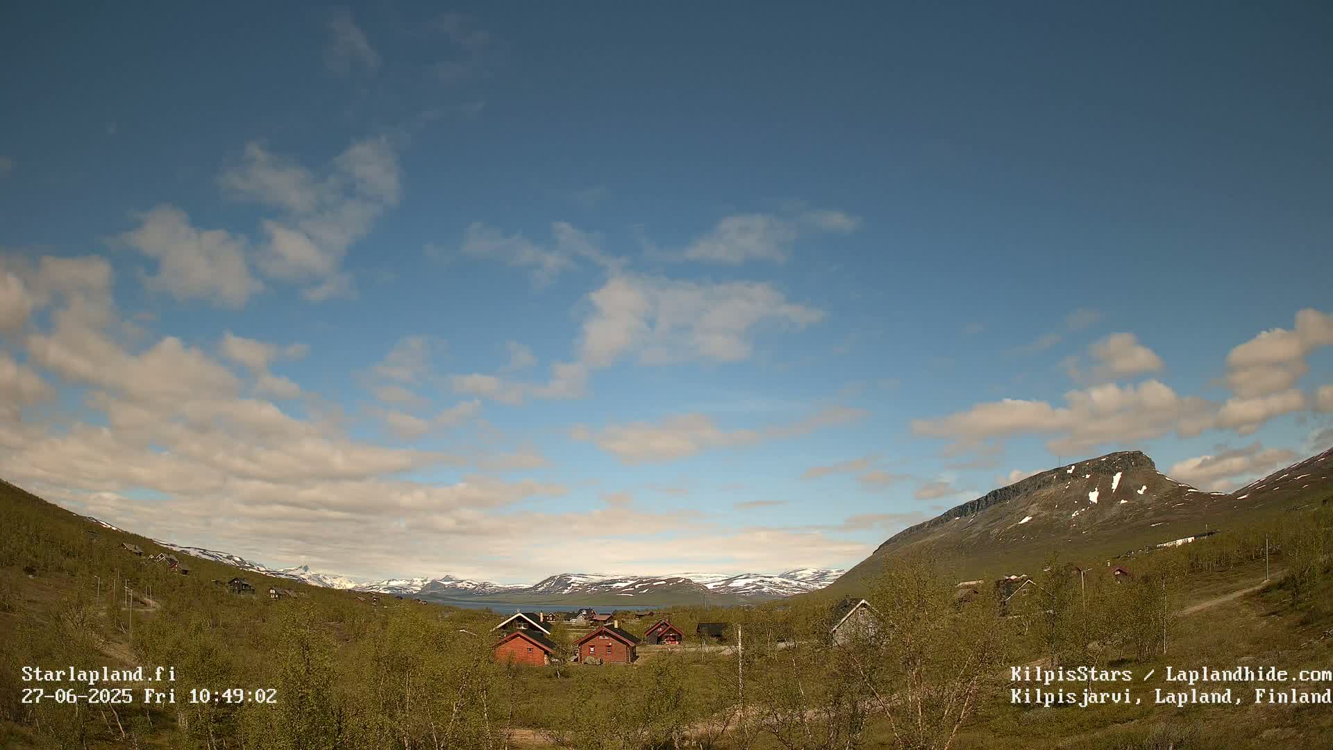 A cluster of small cabins sits in a valley between green hills and snow-capped mountains under a partly cloudy blue sky.