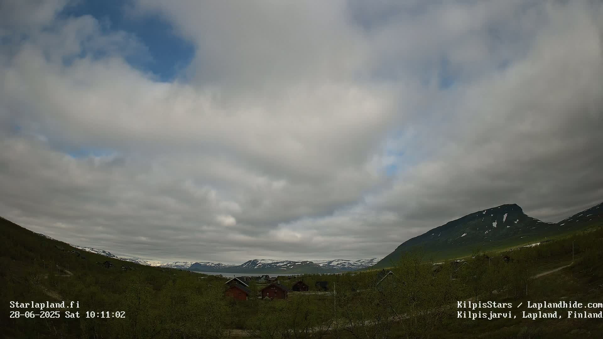 A mostly cloudy sky overlooks a valley of green vegetation and small cabins near a body of water, with snow-capped mountains in the background.