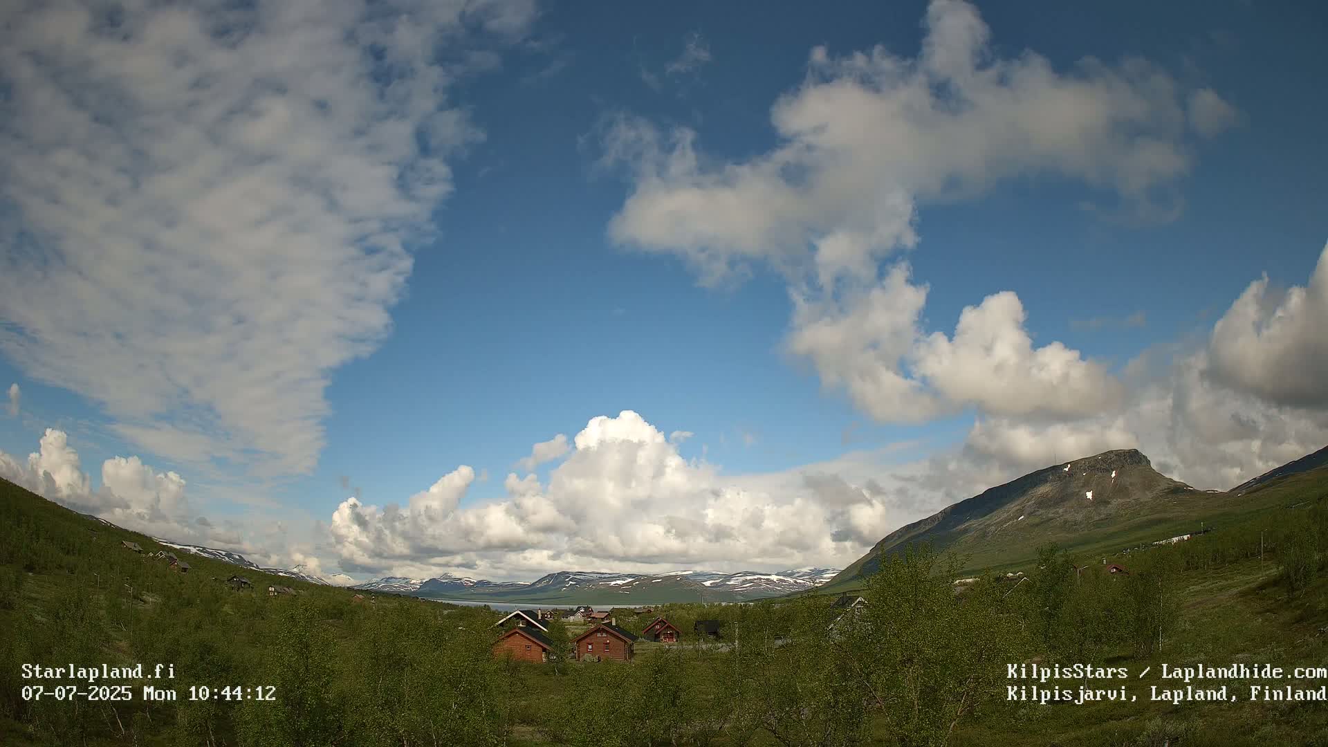 A partly cloudy sky overlooks a valley of green vegetation and several small cabins, with mountains in the background showing patches of snow.