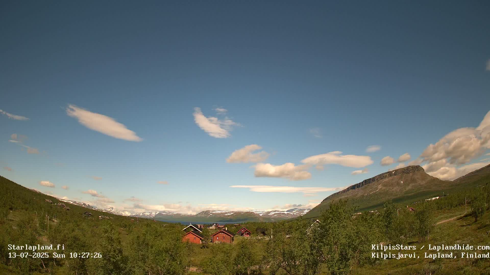 A sunny day with scattered clouds reveals a cluster of red cabins nestled in a valley between green hills and a distant mountain range with patches of snow.