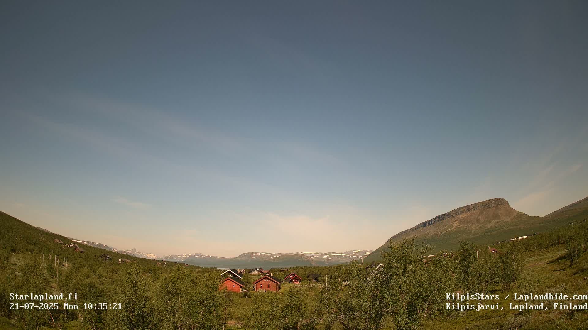 Under a clear, light blue sky, several small red cabins sit nestled in a verdant valley, with mountains visible in the distance.