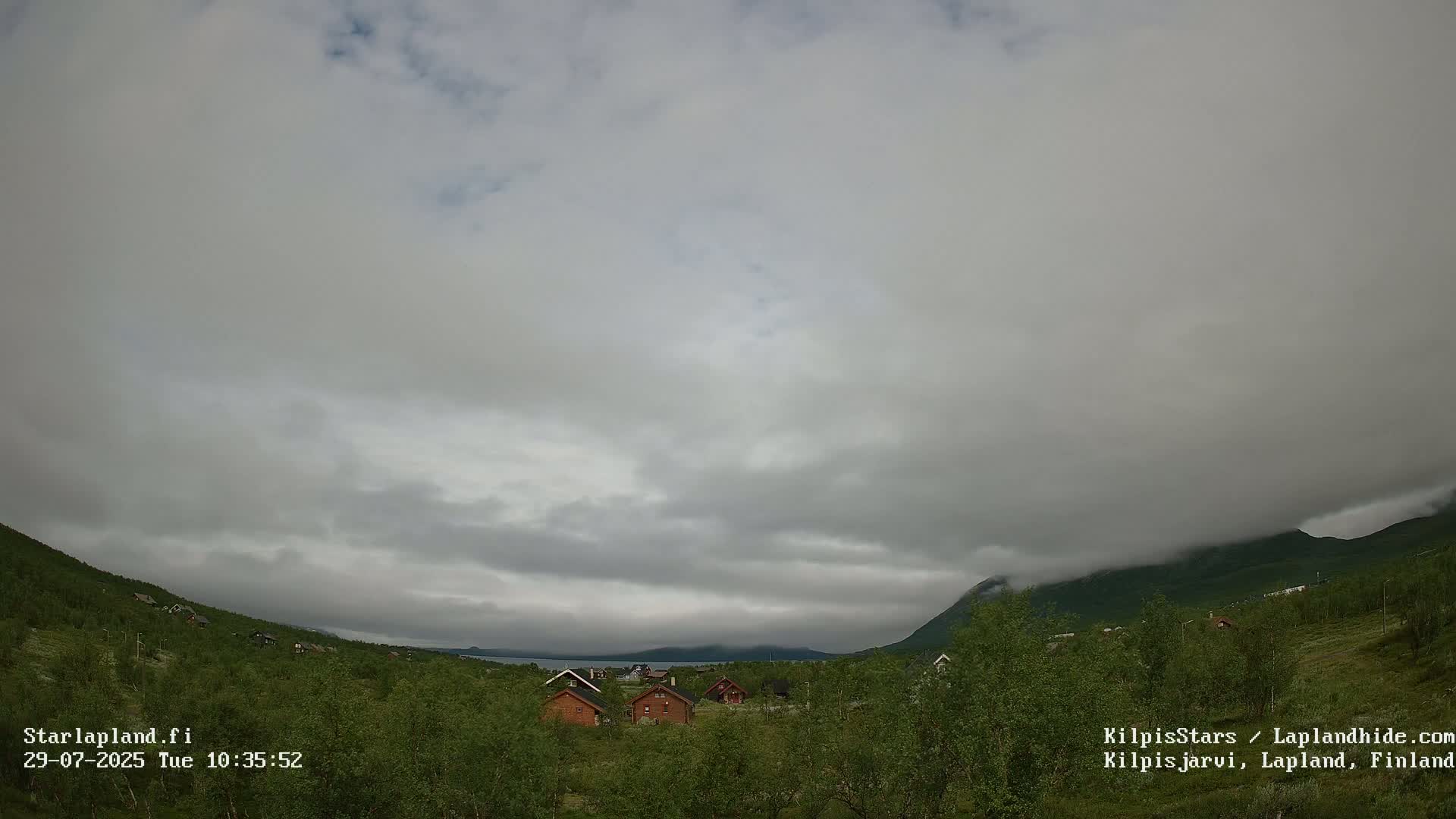 A cloudy sky hangs over a small cluster of houses nestled in a green valley between low mountains.
