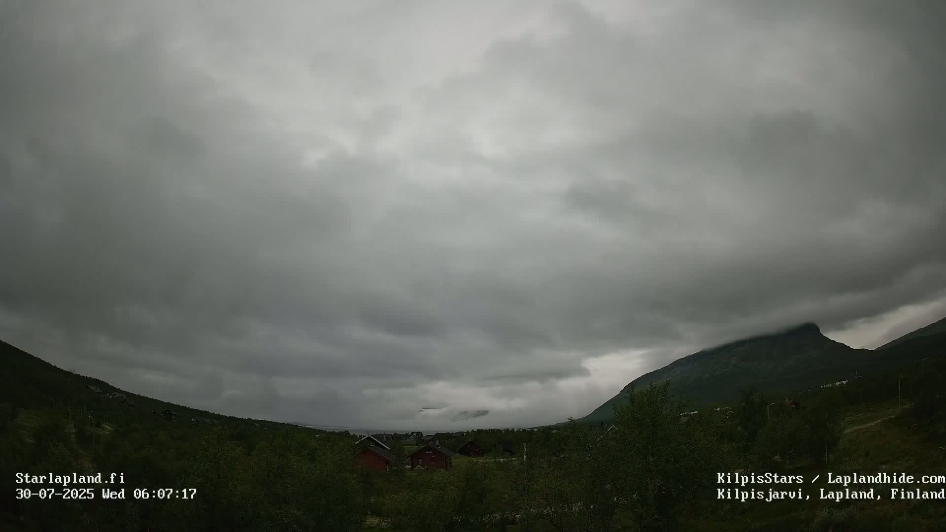 A cloudy sky hangs over a small cluster of houses nestled in a green valley between low mountains.