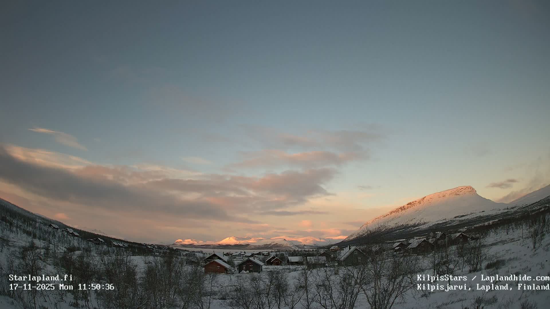 A serene snow-covered village is nestled between rugged snow-capped mountains, bathed in the soft, warm light of a low winter sun under a partly cloudy sky with hints of blue, pink, and orange.