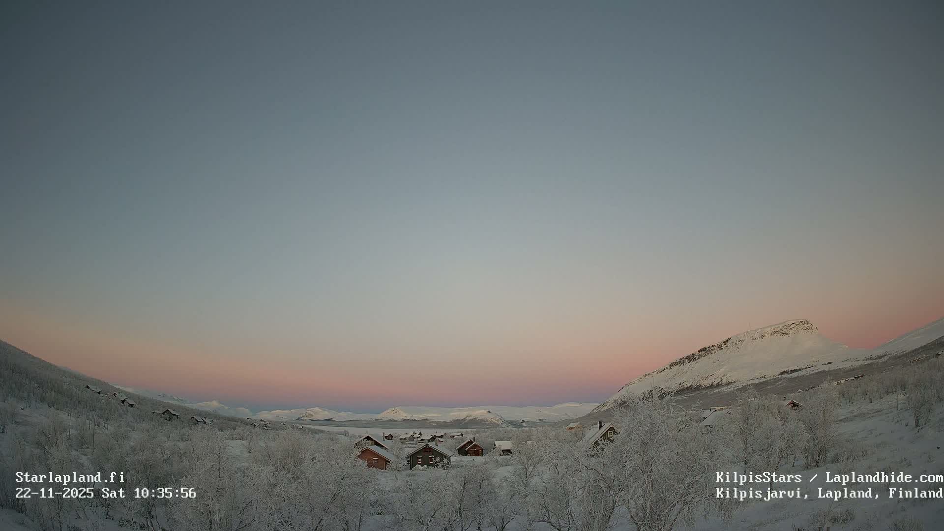 A serene, snow-covered village with frosted trees is nestled in a wide mountain valley under a clear twilight sky transitioning from blue to soft pink, indicating cold winter conditions.