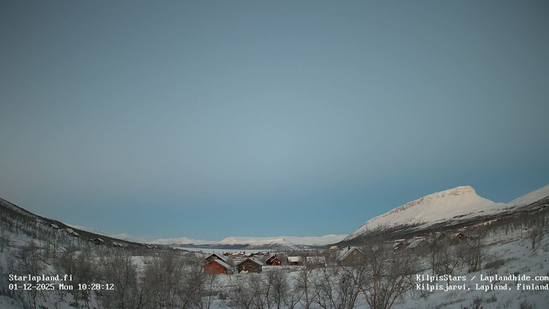 A serene, clear winter day reveals a snow-covered village with scattered houses and bare trees nestled in a valley, flanked by majestic snow-capped mountains under a pale blue sky.