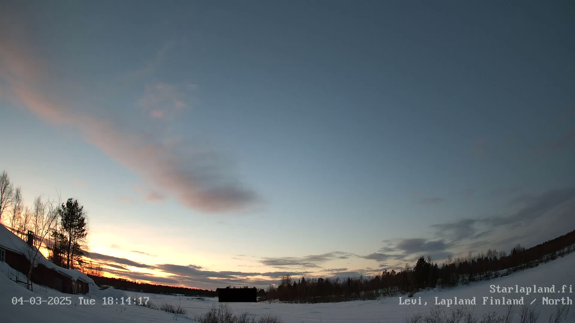 A snow-covered landscape at sunset under a mostly clear sky with a few wispy clouds showing pale pink hues.