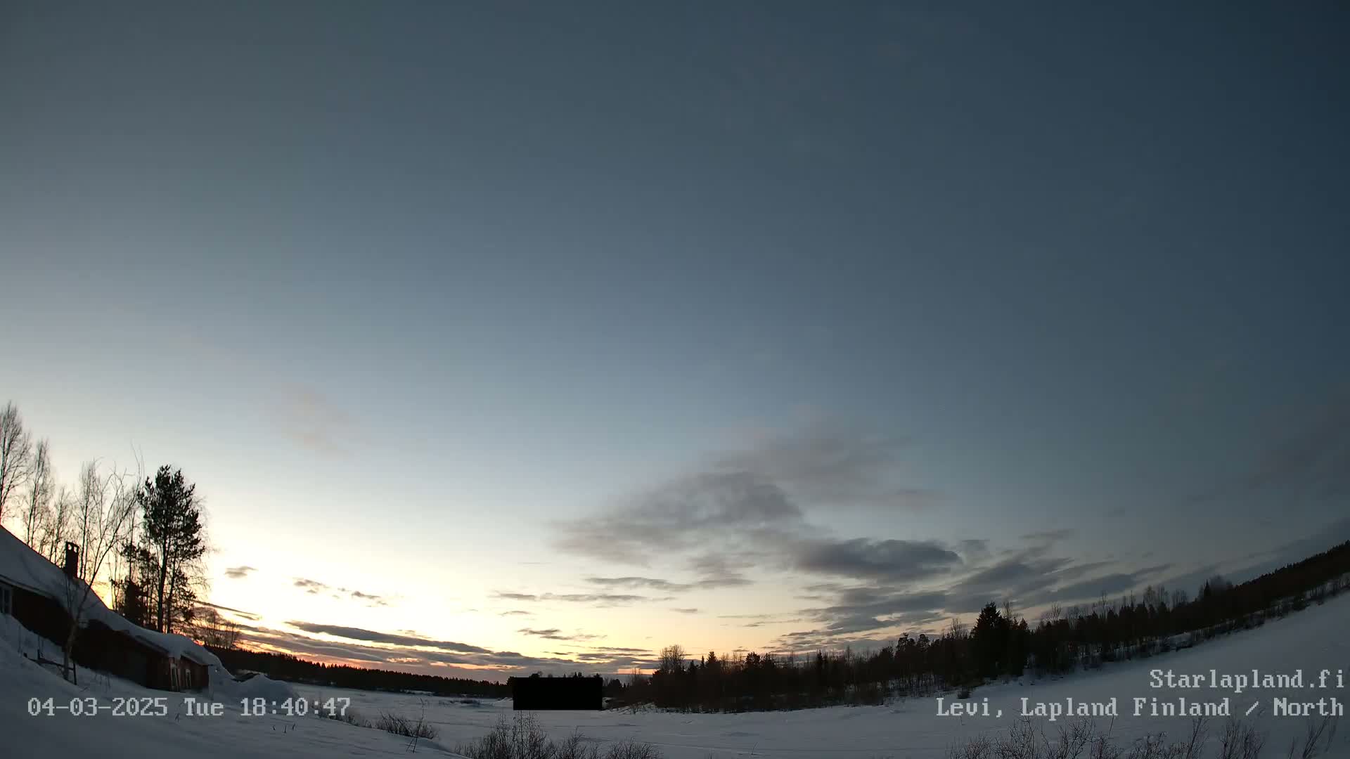 A snow-covered landscape at sunset, under a partly cloudy sky, shows a small building near a treeline.
