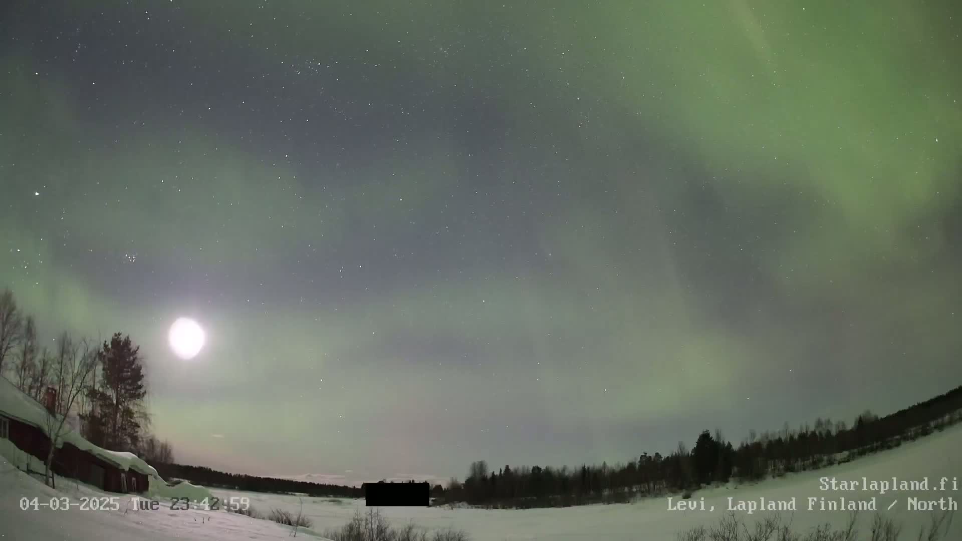 A bright moon and a green aurora borealis illuminate a snow-covered landscape with a dark building and trees under a starry night sky.