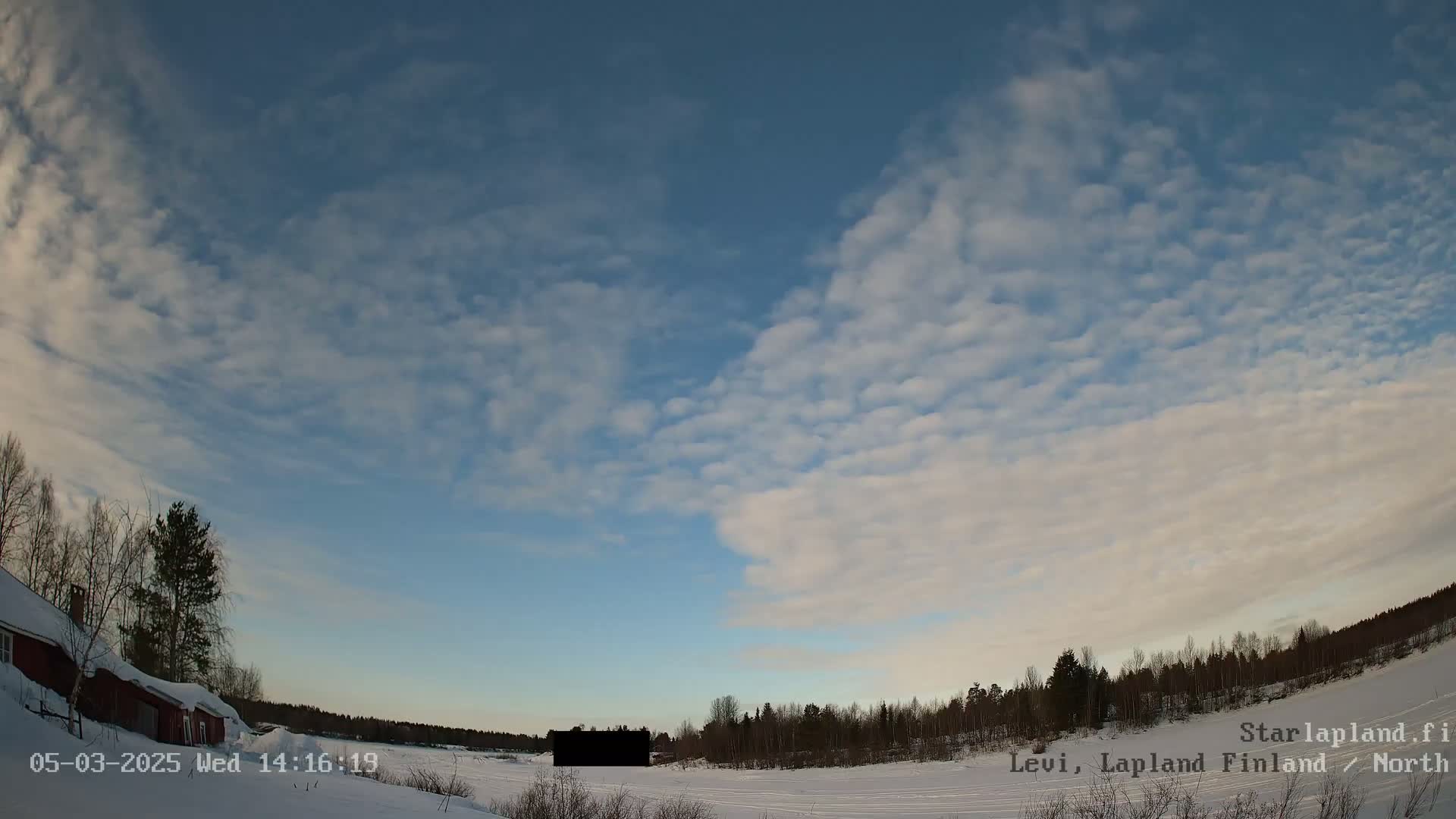 A mostly clear, blue sky with scattered, puffy clouds covers a snowy, flat landscape with a red building and a line of dark trees in the distance.