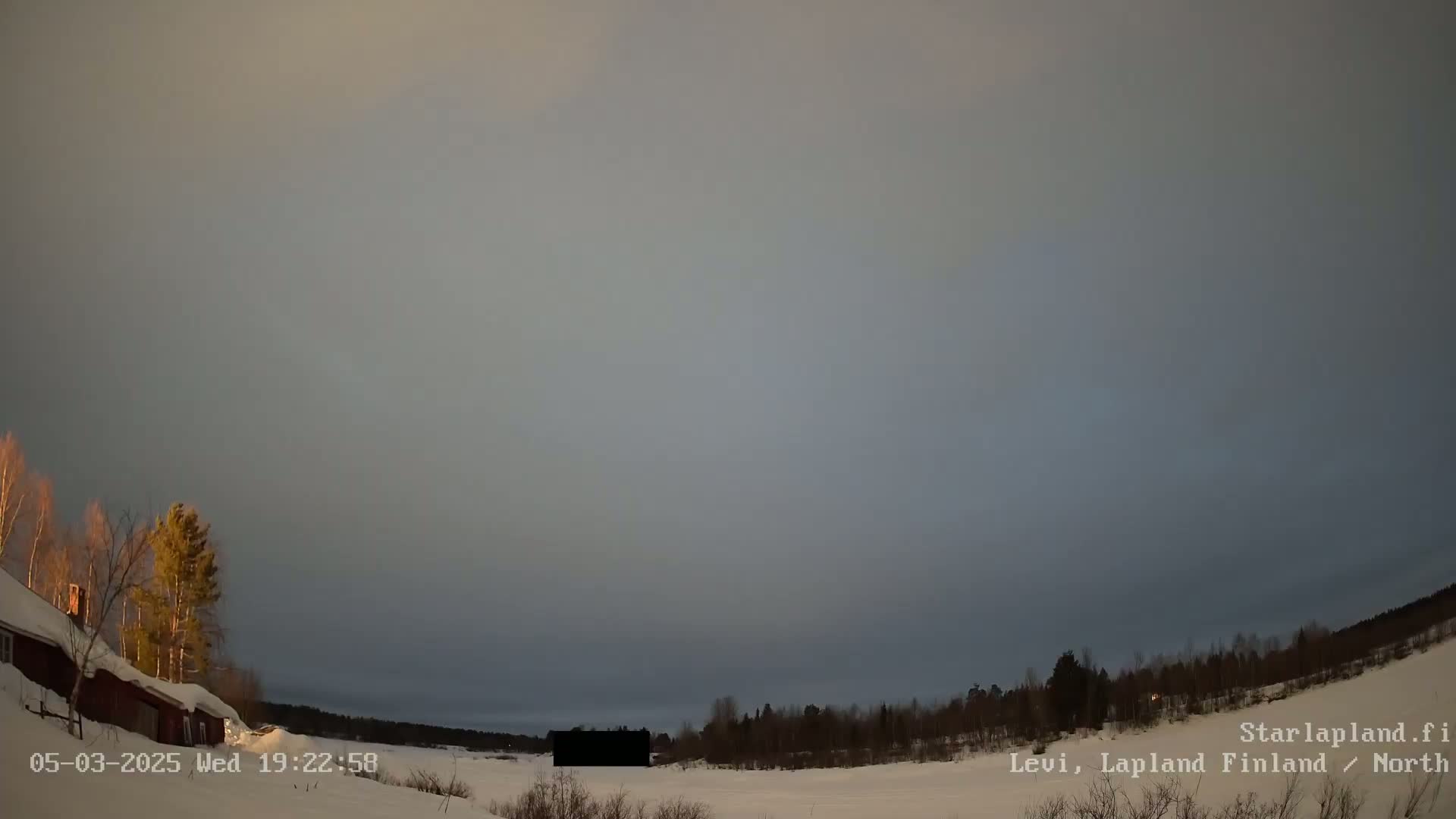 A snow-covered landscape with a few buildings and trees under a mostly cloudy, twilight sky.