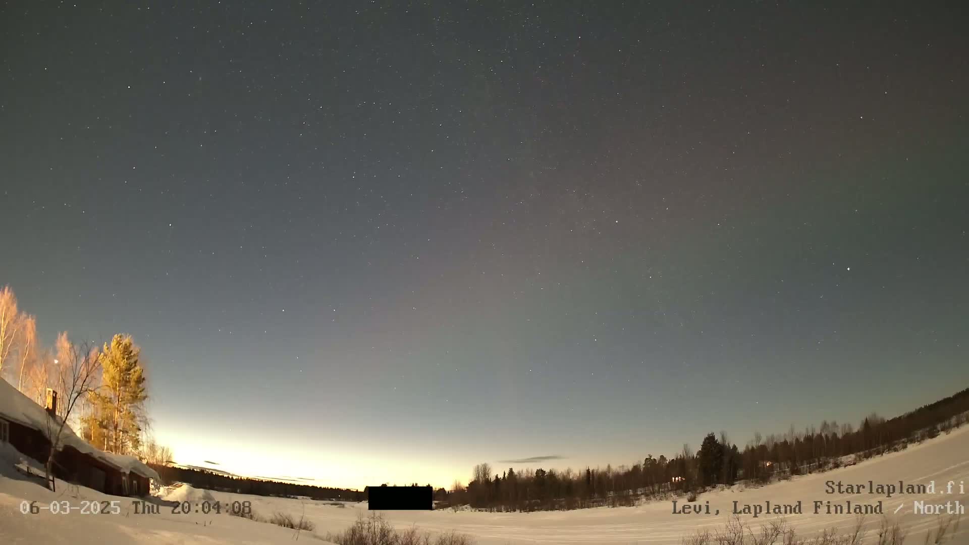 A snow-covered landscape under a night sky filled with stars and a faint aurora borealis, with a dark building and trees in the foreground.