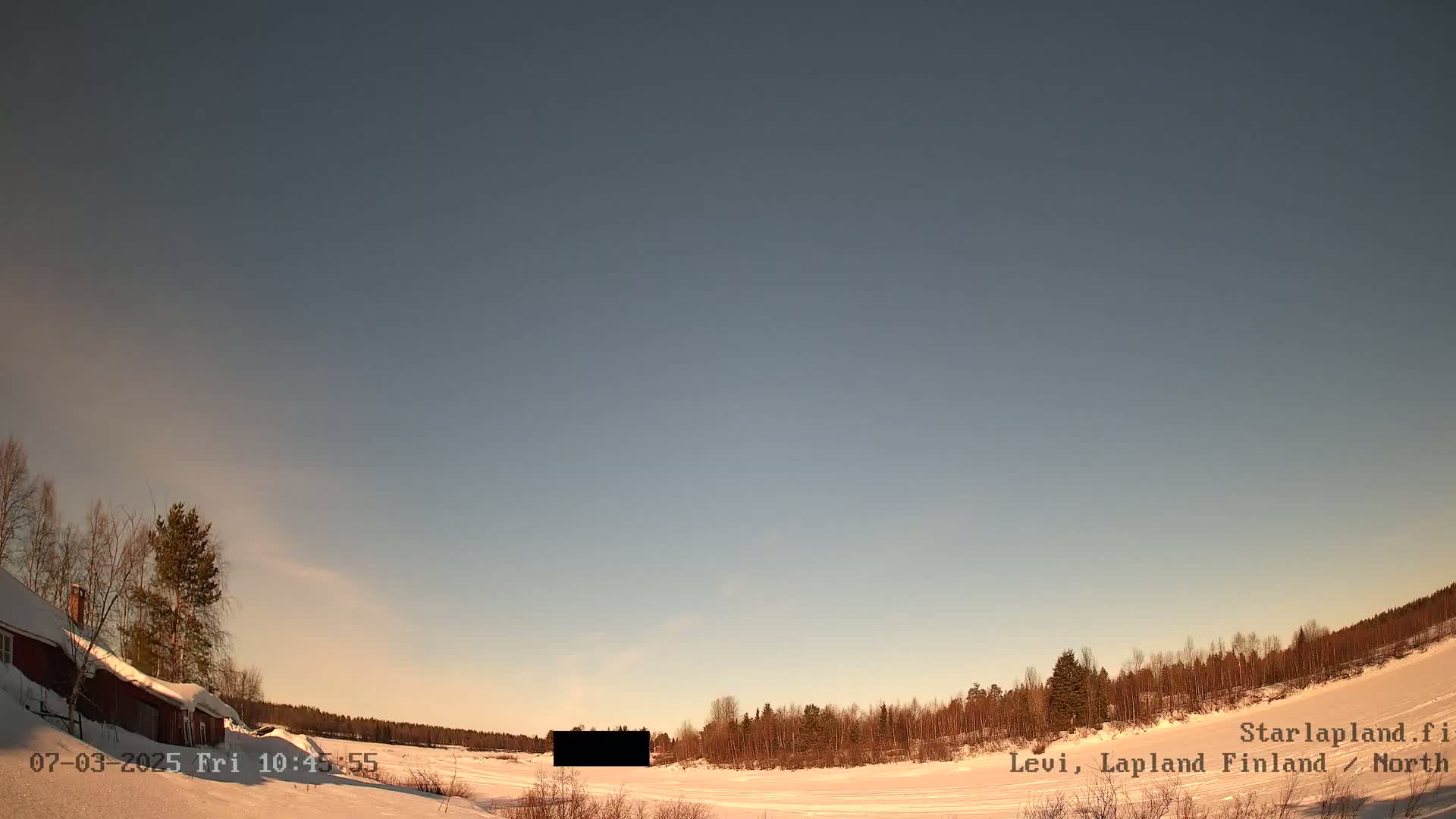 A snow-covered landscape with a red building and leafless trees under a clear, light-blue sky.