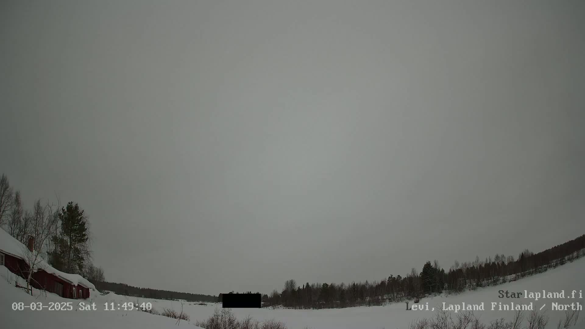 A snow-covered landscape with a dark gray sky and a red building partially visible on the left, featuring leafless trees and a line of evergreens in the background under an overcast sky.
