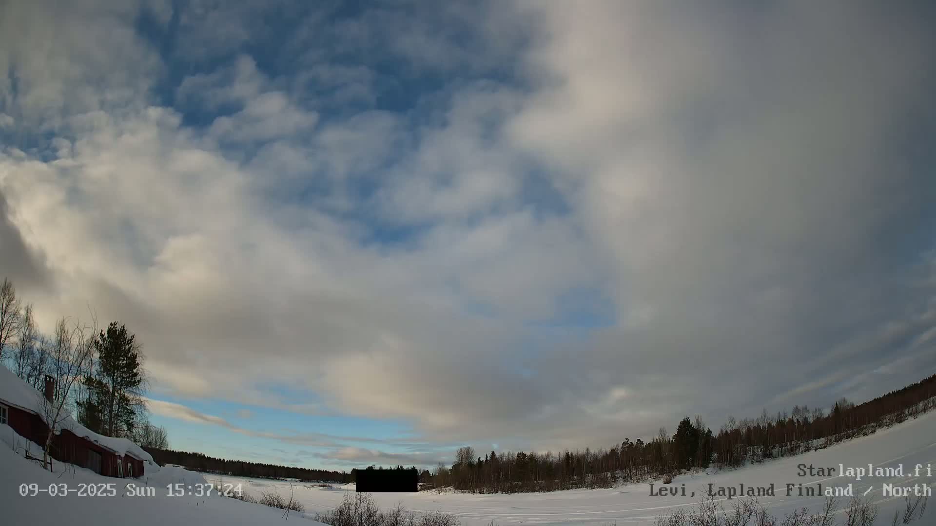 A mostly cloudy, partly sunny sky over a snow-covered landscape with a red building, trees, and a dark rectangular object in the middle distance.