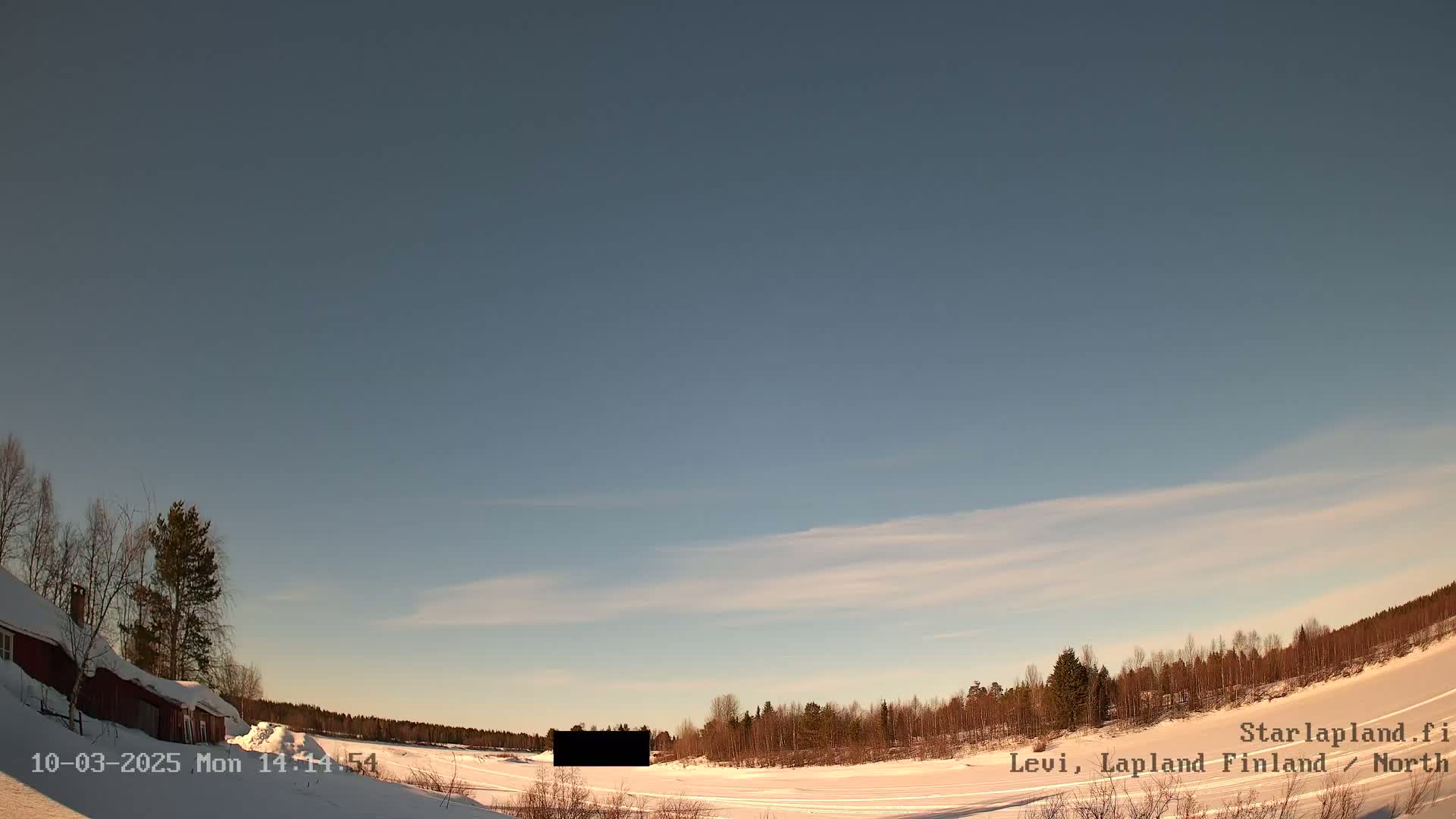 A snow-covered landscape with a red building and leafless trees under a mostly clear, light blue sky.