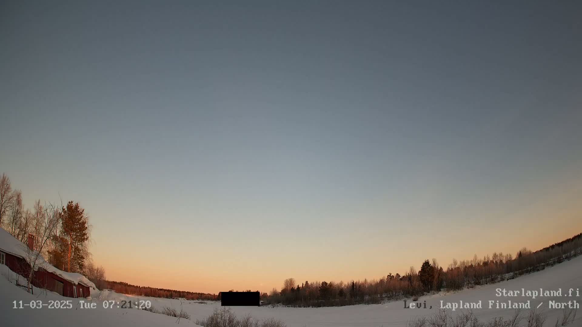 A snow-covered landscape at sunset, featuring a red house and a line of leafless trees under a clear, pale sky.