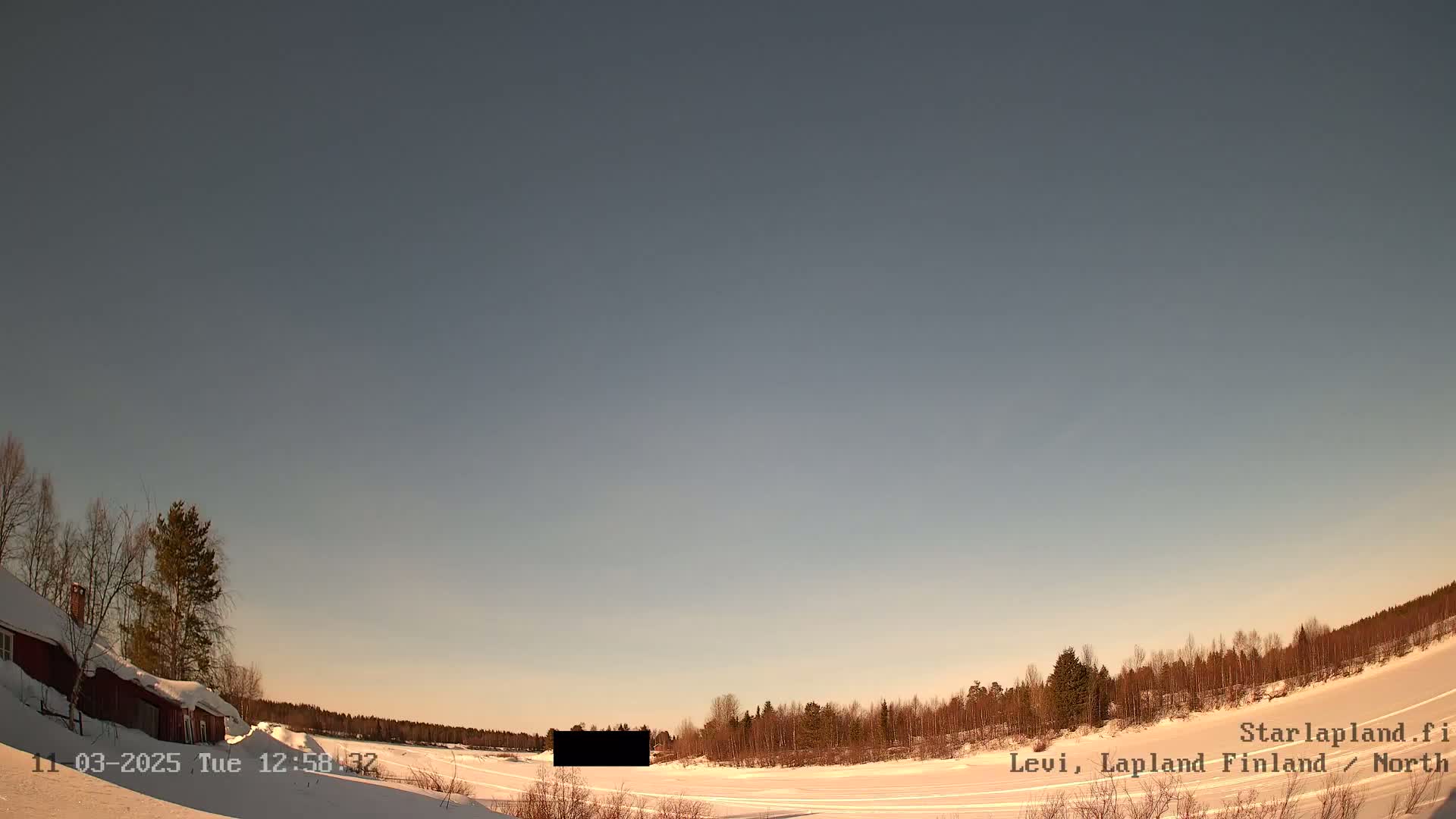 A snow-covered landscape under a clear, pale sky shows a red building to the left and a line of trees in the distance.