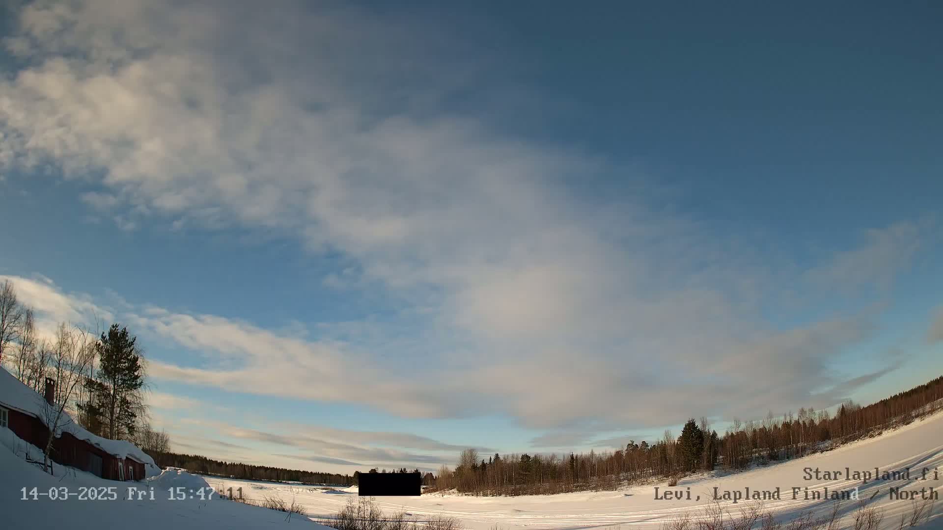 A mostly clear, bright sky with thin clouds stretches over a snowy, flat landscape featuring a red building, leafless trees, and a dark, rectangular object in the mid-ground.