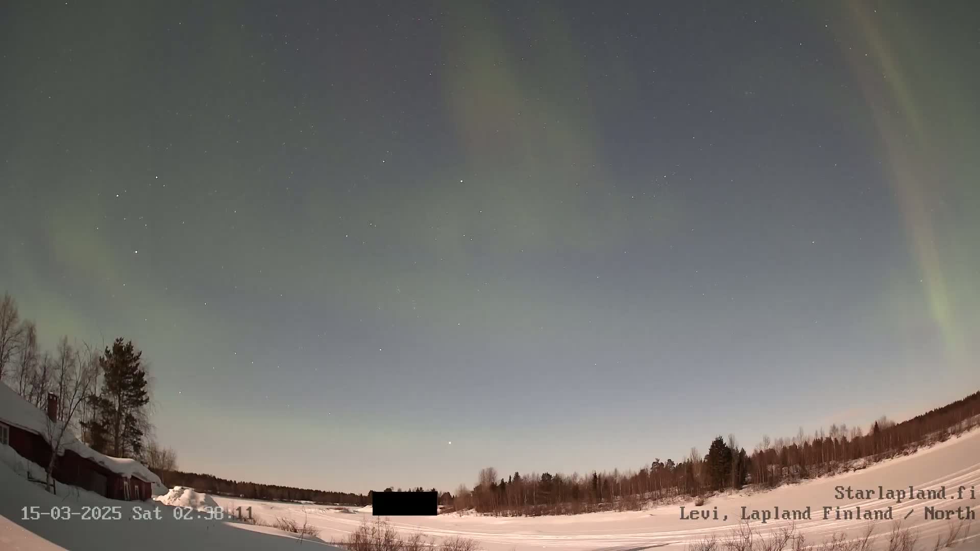A snow-covered landscape at night displays a faint aurora borealis in the sky above a small building and a line of trees.