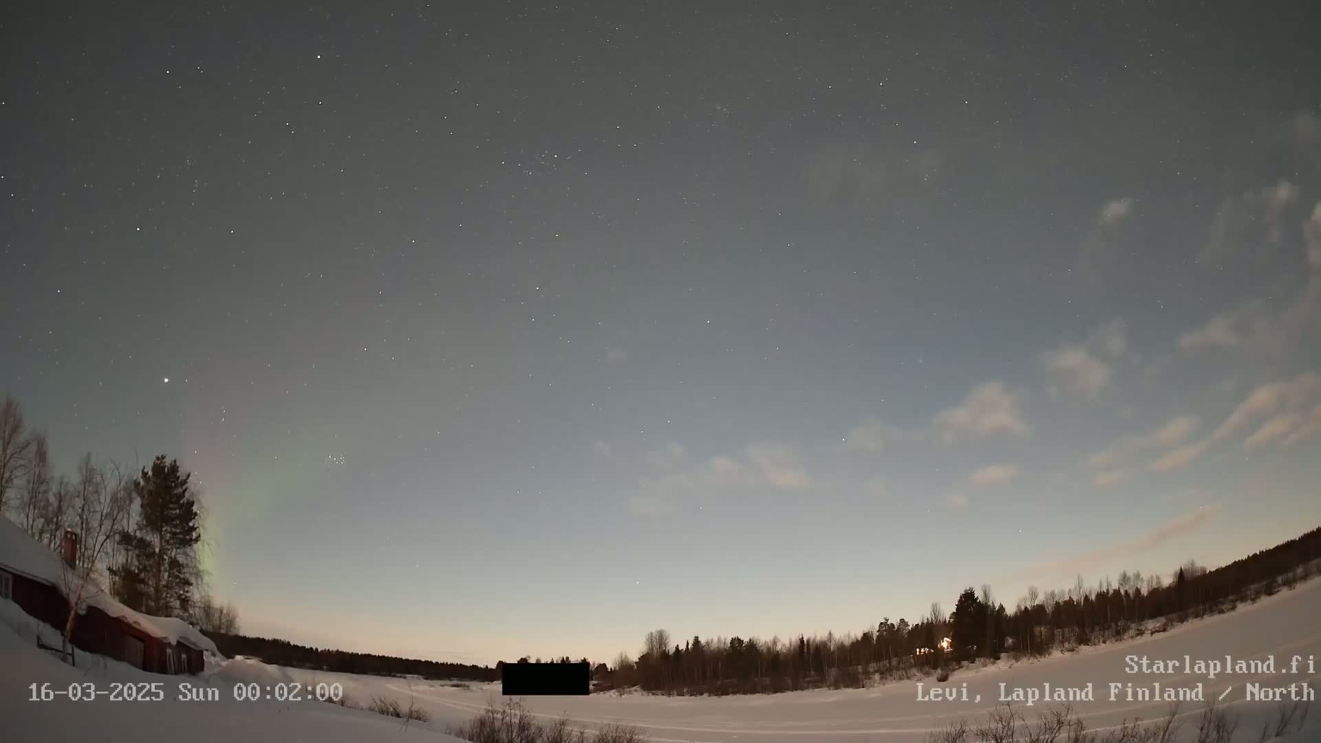 A snow-covered landscape under a night sky with stars and a faint aurora borealis, featuring a few buildings and trees in the foreground.