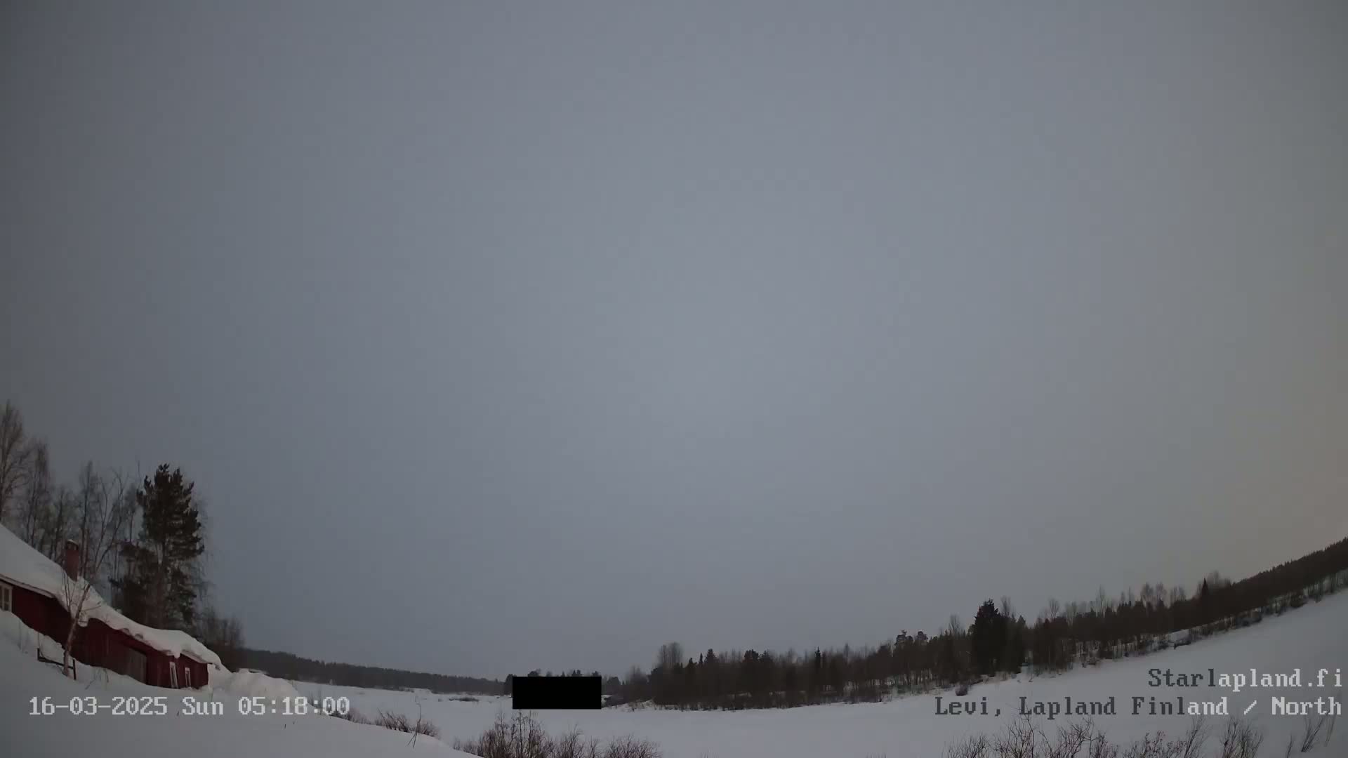 A snow-covered landscape under an overcast sky shows a red building to the left and a line of dark trees in the distance.