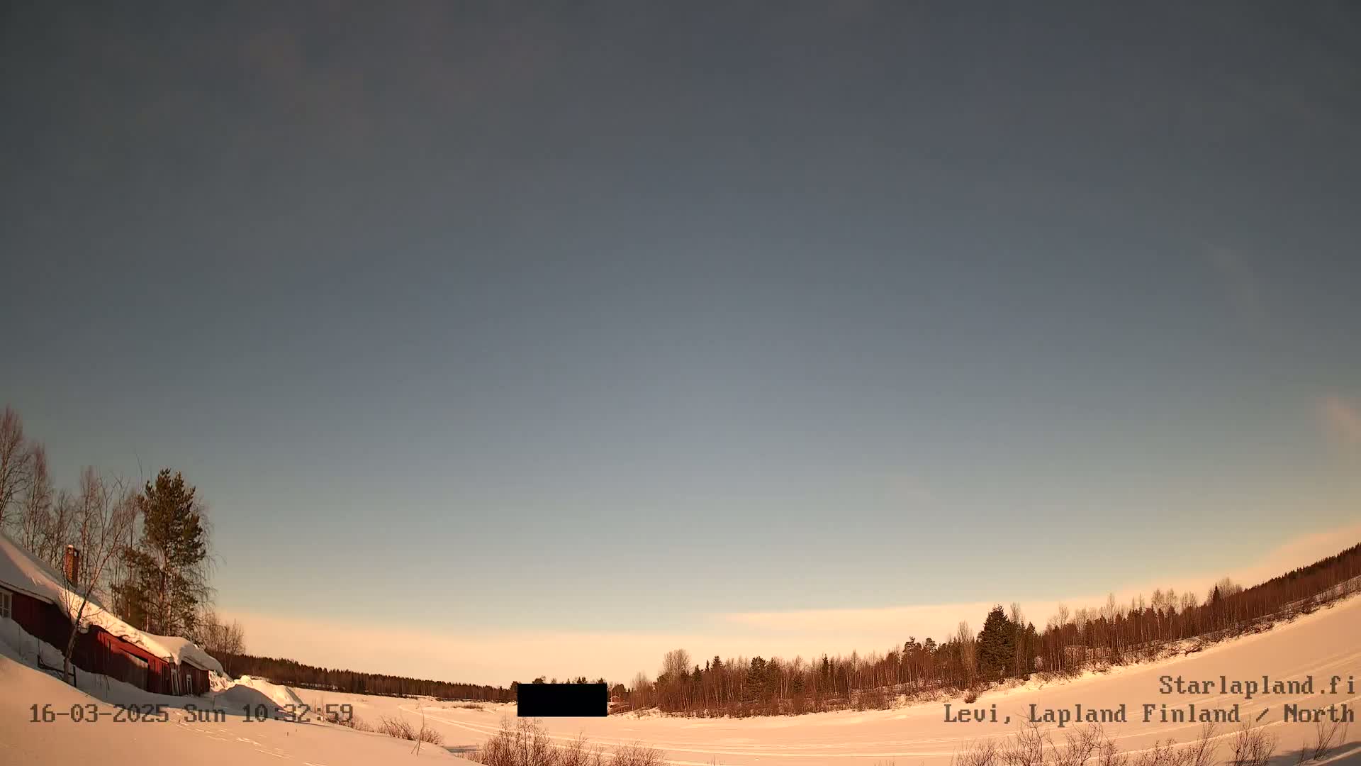 A snow-covered landscape with a red building and a line of trees under a mostly clear, pale sky.