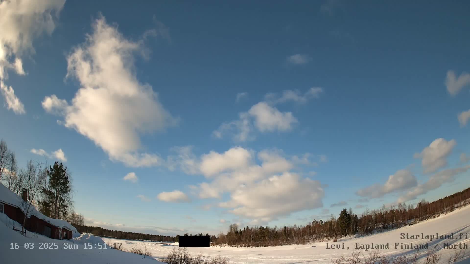 A partly cloudy blue sky overlooks a snow-covered landscape with sparse trees and a red building in the left corner.