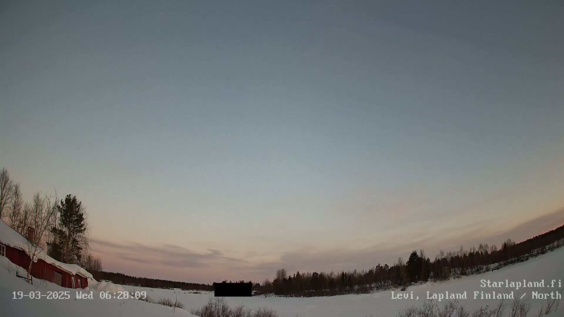 A red building is partially visible on the left, nestled in a snowy landscape with a line of trees in the distance under a twilight sky.