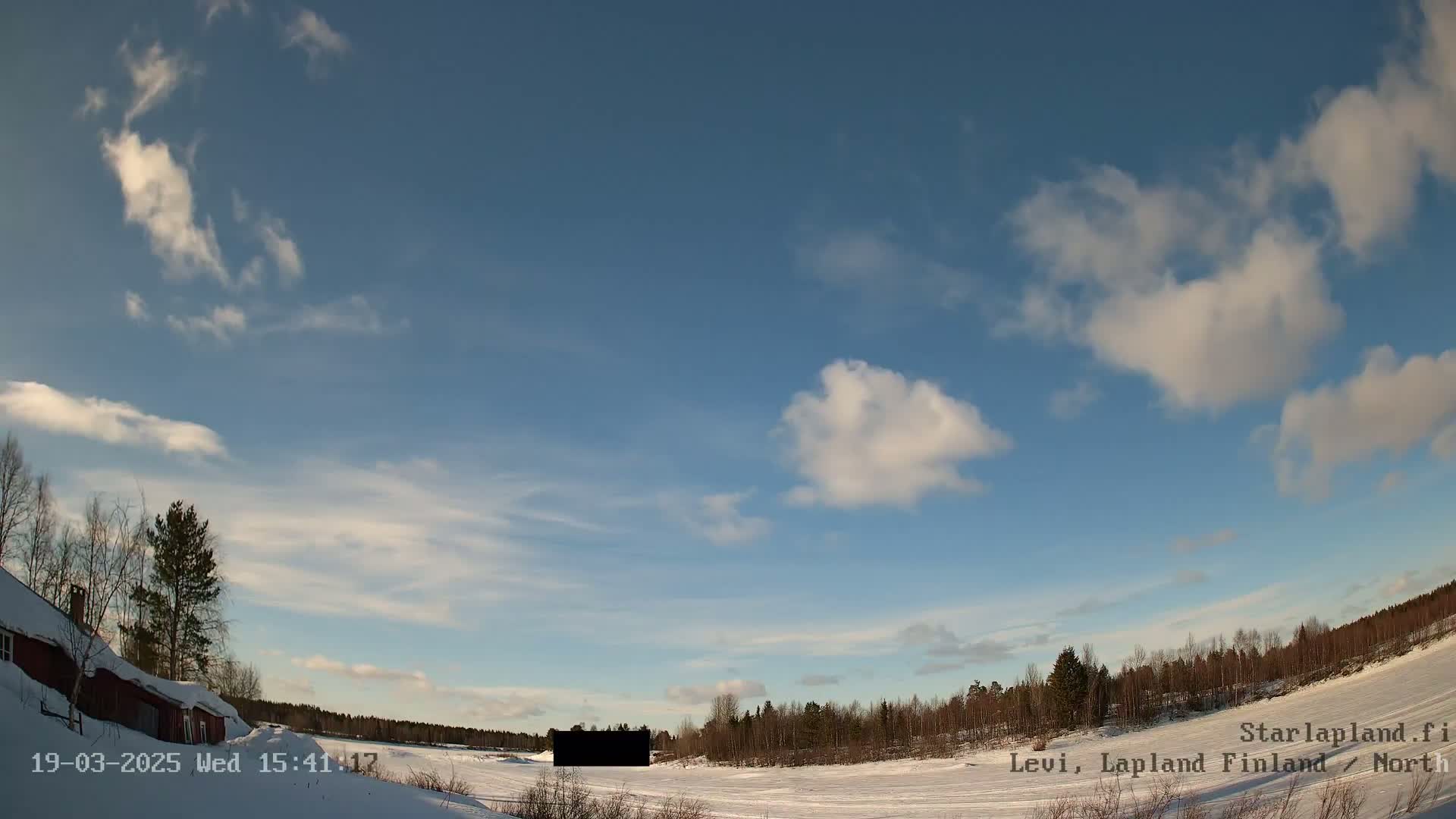 A partly cloudy, sunny winter day with snow covering the ground and trees, showing a red building to the left and a forest in the distance.
