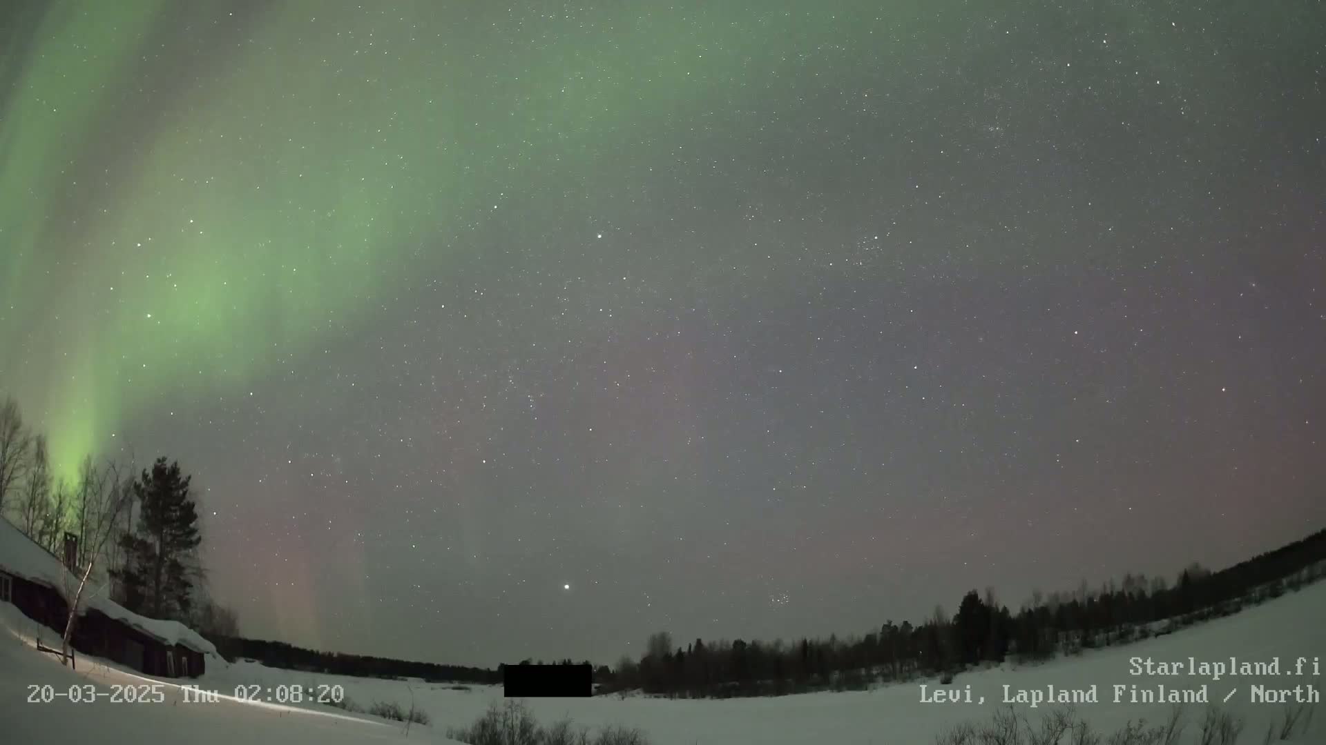 A vibrant green aurora borealis illuminates a night sky over a snow-covered landscape with a dark building and trees visible in the foreground.