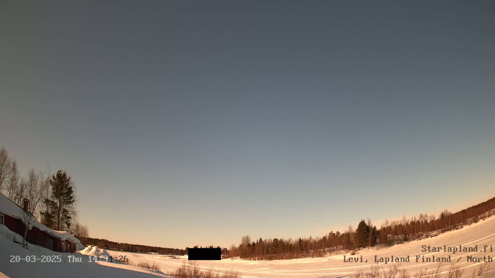 A snow-covered landscape with a red building and bare trees under a clear, pale sky.