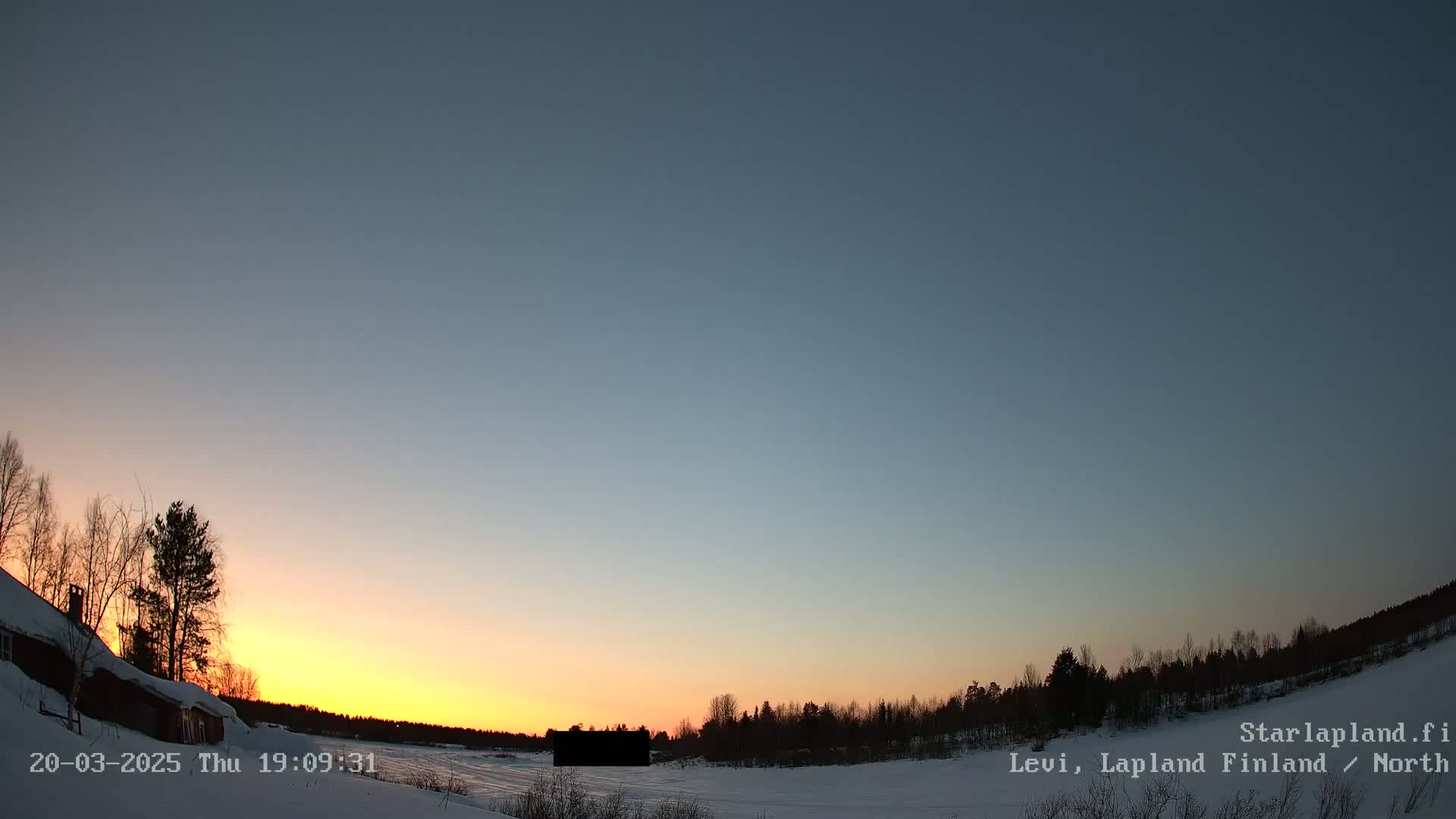 A snow-covered landscape at sunset, showing a partially visible building to the left, and a line of dark trees on the horizon under a clear, twilight sky.