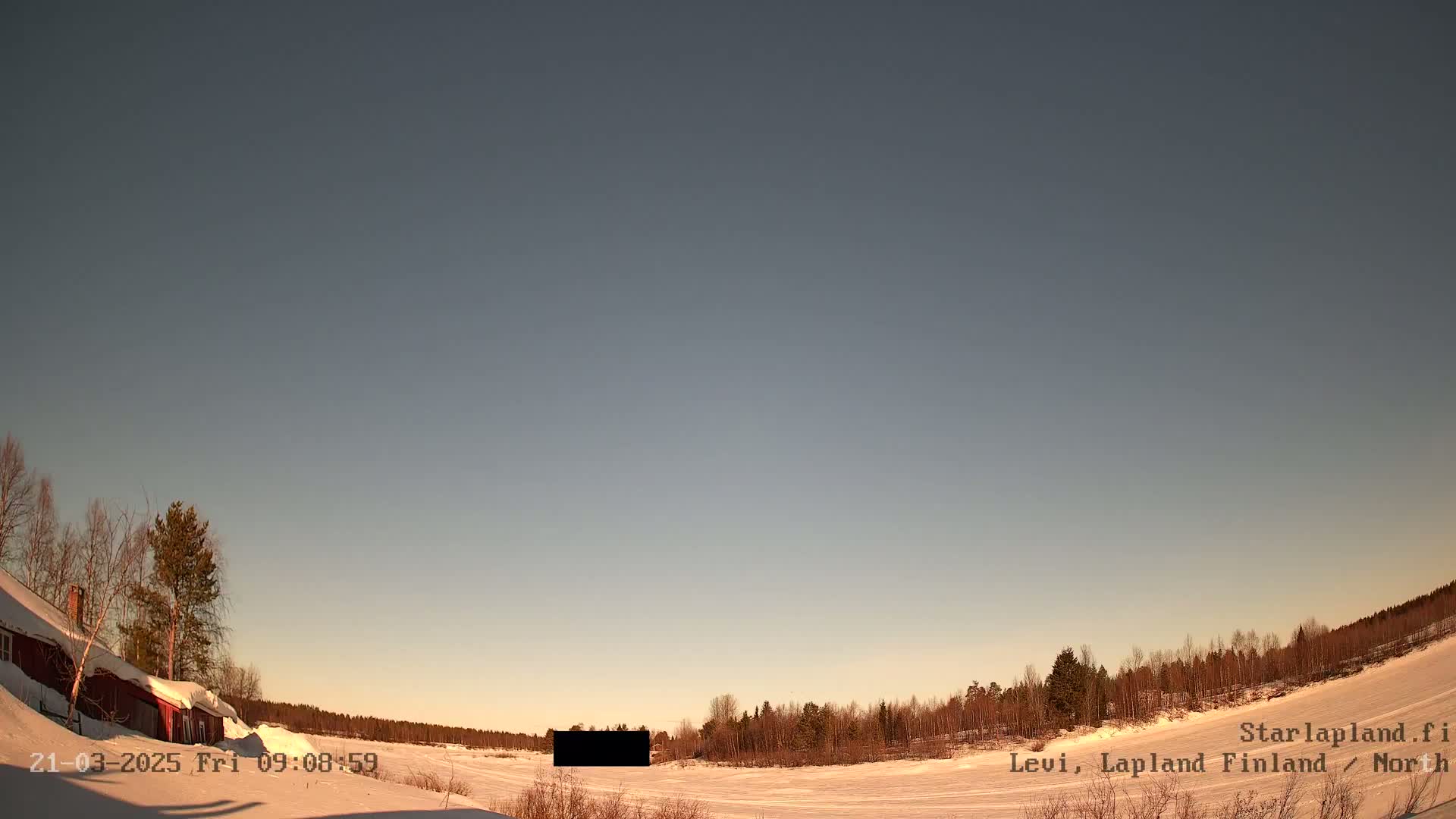 A snow-covered landscape with a red building and bare trees under a clear, light-blue sky.